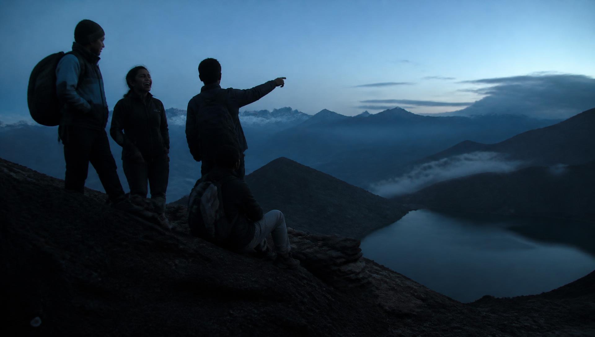 Young Indian travellers with backpacks on a ridge above a Himalayan lake at blue hour, misty peaks behind them